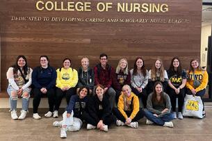 A group of students sit under a sign that says College of Nursing at the University of Wisconsin–Oshkosh